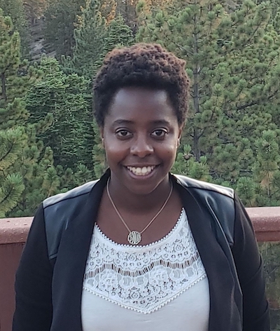 Photo of Sandra Musaka smiling and standing in front of some greenery. 